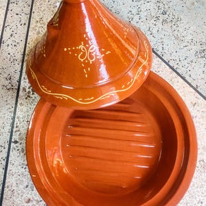 Puede incluir: Una tagine de terracota con una tapa que presenta un diseño decorativo en amarillo. La tagine tiene una base acanalada.