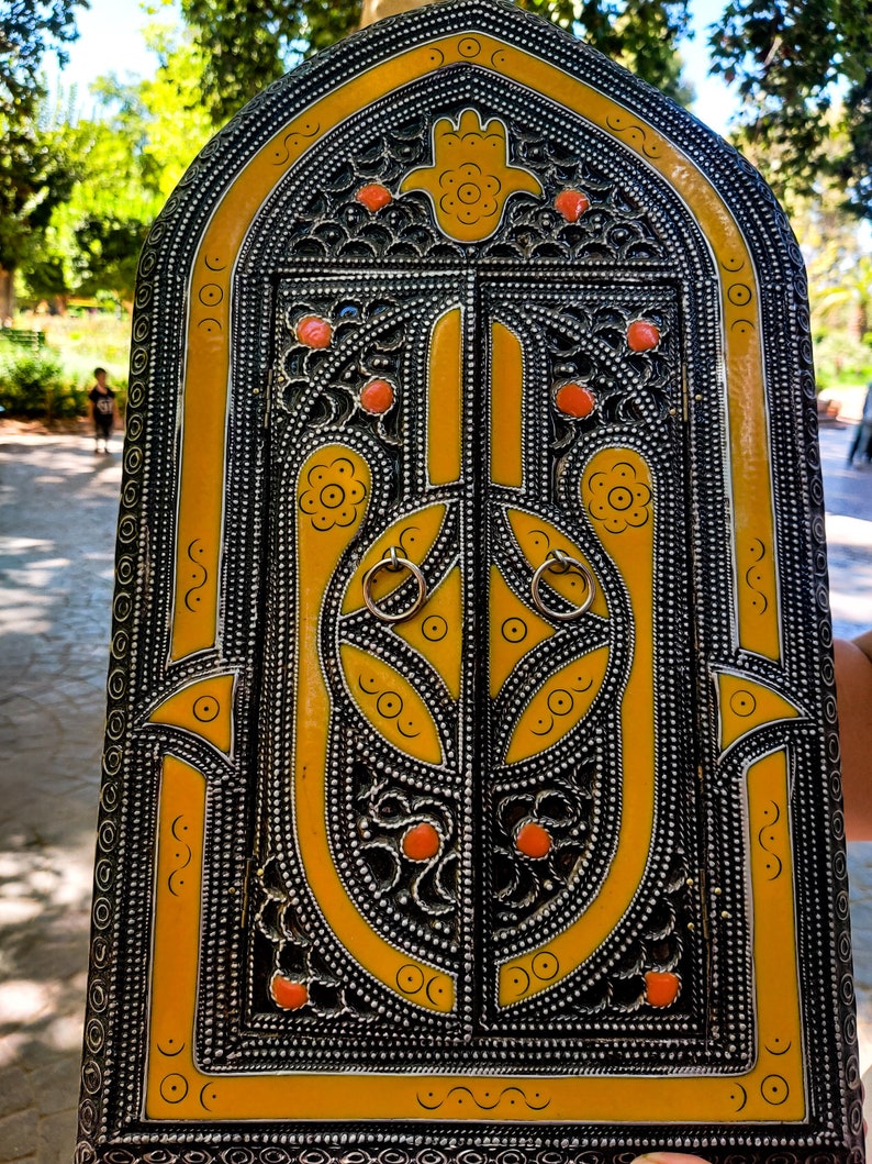 May include: A yellow and black metal decorative box with a carved design of a hamsa hand. The box has two doors and is decorated with small circles and floral patterns.