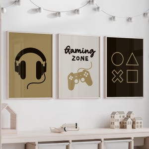 May include: Three framed prints with a gaming theme. The first print has a pair of black headphones on a tan background. The second print has the text "gaming zone" in black on a tan background. The third print has a black background with a white circle, triangle, square, and X.