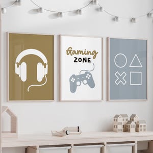 Pode incluir: Três impressões emolduradas com um tema de jogos. A primeira impressão tem um fundo dourado com uma silhueta branca de fones de ouvido. A segunda impressão tem um fundo branco com o texto "gaming zone" em preto e um controlador de videogame cinza. A terceira impressão tem um fundo cinza com contornos brancos de um círculo, um triângulo, uma cruz e um quadrado.