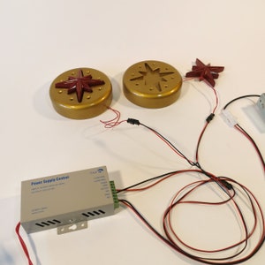Peut inclure: Gros plan sur une unité de contrôle d'alimentation avec des fils connectés à deux luminaires en forme d'étoile dorés. L'unité d'alimentation a une étiquette qui indique "Power Supply Control" et a le texte "INPUT AC100V-240V 50/60HZ" et "OUTPUT DC12V 5A".