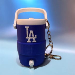 Puede incluir: Un enfriador azul y blanco en miniatura con el logotipo "LA" de los Dodgers de Los Ángeles. El enfriador tiene un asa blanca, un grifo y un llavero plateado. El enfriador mide aproximadamente 10 cm de alto.