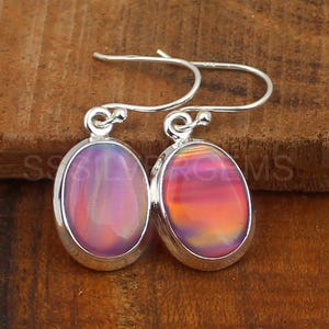 May include: A pair of silver drop earrings with oval-shaped, iridescent opal stones. The opals display a blend of pink, orange, and purple hues, set in a polished silver frame. The earrings are displayed on a wooden surface.