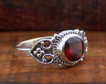 Puede incluir: Un anillo de plata con una gema grande, redonda y de color rojo intenso. El anillo tiene un diseño ornamentado con remolinos decorativos y pequeños detalles de cuentas alrededor del engaste de la gema.