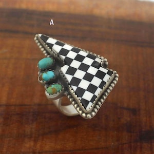 Pode incluir: Um anel de prata com um design triangular com um padrão xadrez preto e branco. O anel é adornado com três pedras turquesas e uma borda com contas. O anel está sobre um fundo de madeira.