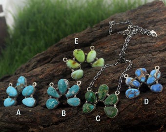 Op de afbeelding: Een collectie turquoise en groene stenen sieraden, waaronder kettingen en hangers. De stenen zijn gezet in zilver en gerangschikt in clusters. De achtergrond is een donker houten oppervlak met groen gras.