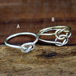 May include: Two rings, one silver and one gold, each featuring a knot design. The silver ring is a single band with a knot at the center. The gold ring has a gold band and a silver knot.