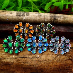 May include: A collection of gemstone stud earrings in various colors, including orange, green, and blue. Each pair features a cluster of oval-shaped stones set in a silver-toned metal. The earrings are displayed on a wooden surface.