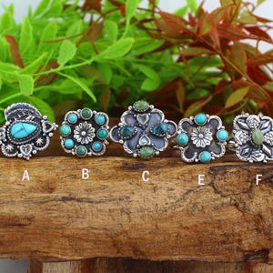 May include: A collection of six silver-toned rings, each adorned with turquoise and green gemstones. The rings feature various designs, including floral and geometric patterns. The gemstones are set in intricate metalwork, showcasing a Southwestern or bohemian style.