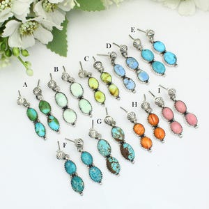 May include: A collection of nine pairs of drop earrings labeled A through I, each featuring two oval-shaped stones set in silver with a scalloped top detail. The stones vary in color, including shades of green, blue, yellow, orange, and pink.