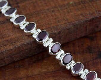 May include: A silver bracelet featuring oval-cut, deep red gemstones. The gemstones are set in individual silver links, creating a continuous chain. The bracelet is displayed on a wooden surface, highlighting the contrast between the silver and the gemstones.