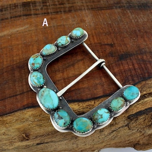 May include: A silver belt buckle featuring turquoise stones. The buckle has a rectangular shape with a curved base and a silver bar for the belt. The turquoise stones are oval and set around the edges of the buckle. The buckle sits on a wooden surface.