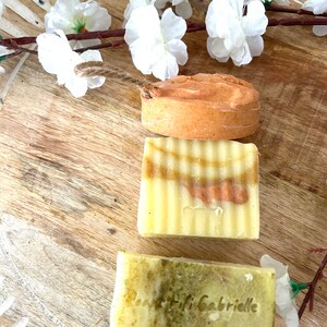 May include: Three handmade soaps in shades of yellow, orange, and green are arranged on a wooden surface, decorated with white flowers. The soaps have unique textures and colours, with one bearing the inscription "Bonne Fifi Gabrielle".