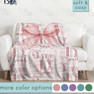 May include: A white blanket with the name "Sarah" in a repeating pattern and a large pink bow. The blanket is draped over a white couch. The text "soft & cozy" is in the upper right corner. The words "more color options" are at the bottom.