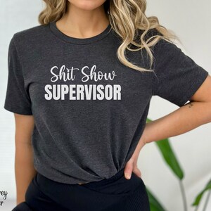 May include: Dark grey heather t-shirt with the text "Shit Show Supervisor" in white lettering.