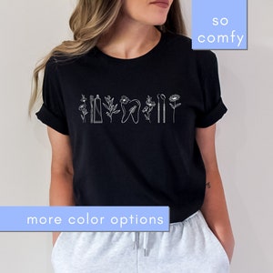 Peut inclure: T-shirt noir avec un motif blanc d'outils dentaires et de fleurs. Le texte "so comfy" est dans un encadré bleu au-dessus du t-shirt et "more color options" est dans un encadré bleu en dessous du t-shirt.