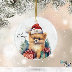 May include: A white ceramic Christmas ornament featuring a small, brown and white Pomeranian dog wearing a red Santa hat with the year 2024 on it. The dog is holding a red and white gift box and a green gift box. The ornament is personalized with the name "Chase".