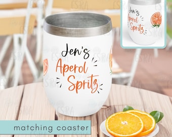 Peut inclure: Un gobelet blanc avec un couvercle argenté et un motif qui dit "Jen's Aperol Spritz" avec des tranches d'orange et des feuilles vertes. Le gobelet est posé sur une table en bois avec un dessous de verre assorti.