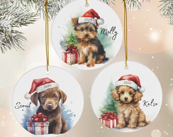 May include: Three round ceramic Christmas ornaments featuring watercolor illustrations of dogs wearing Santa hats. The ornaments are white with gold string hangers. The first ornament features a Yorkshire Terrier with the name "Molly" and the year "2024". The second ornament features a Labrador Retriever with the name "Scout" and the year "2024". The third ornament features a Goldendoodle with the name "Kelso" and the year "2024".