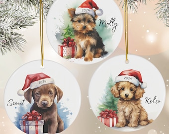 May include: Three round ceramic Christmas ornaments featuring watercolor illustrations of dogs wearing Santa hats. The ornaments are white with a gold string for hanging. The first ornament features a Yorkshire Terrier with the name "Molly" and the year "2024". The second ornament features a Labrador Retriever with the name "Scout" and the year "2024". The third ornament features a  poodle with the name "Kelso" and the year "2024".