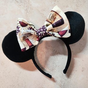 May include: Black mouse ears headband with a fabric bow featuring a wine theme. The bow has a white background with a red wine bottle, grapes, and the text "Chateau Wine" and "Vins Fins Chevrain".