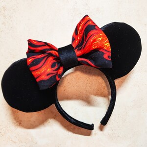 May include: Black mouse ear headband with a red and yellow flame-patterned bow. The ears are round and made of black velvet. The bow is centered on the headband, adding a touch of flair. The headband is a fun accessory.