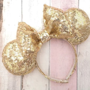 May include: Gold sequin Minnie Mouse ears headband with a large bow. The headband is white.