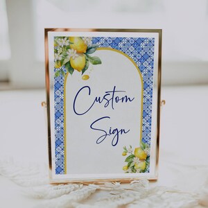 May include: A gold-framed sign with a white background and blue and white patterned border. The sign features a watercolor illustration of lemons and flowers. The text "Custom Sign" is written in blue cursive.