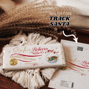 May include: A white chocolate bar with gold lettering reading "Sophie Believes", a red ribbon, and a Christmas ornament illustration. The bar is labelled "Express Delivery" and "25th December". The image also includes a card with a QR code and the text "Track Santa".