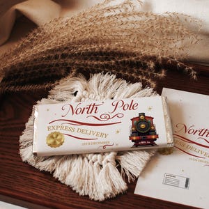 May include: A chocolate bar with a festive "North Pole Express Delivery" design, featuring a vintage train illustration. The bar rests on a white macrame coaster and a matching card, all on a wooden tray.