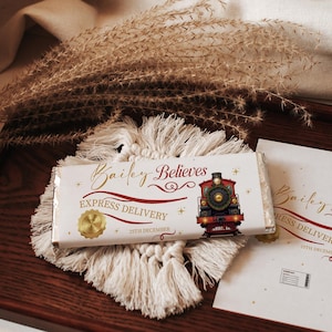 Pode incluir: Uma barra de chocolate branco com o texto "Bailey Believes" e uma ilustração de comboio vermelho e dourado. A barra está sobre uma base de macramé e uma bandeja de madeira. As palavras "Express Delivery" e "25th December" também são visíveis.