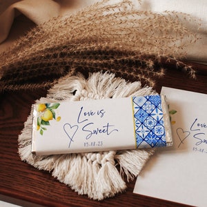 Könnte beinhalten: Eine weiße Schokoladentafel-Verpackung mit einem blauen und weißen gemusterten Rand. Die Verpackung zeigt eine Aquarell-Illustration von Zitronen und grünen Blättern. Der Text "Love is Sweet" ist in blauer Kursivschrift geschrieben, darunter das Datum "05-08-23".