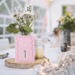 May include: A pink table number card with white text that says "Table 1" in a decorative font. The card is sitting on a wooden slice with a white tablecloth and a jar of white flowers in the background.