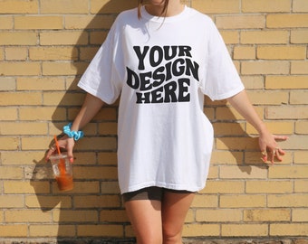 May include: A white oversized t-shirt with the text "YOUR DESIGN HERE" in black. The shirt is being worn by a person standing in front of a brick wall. The person is wearing white sneakers.