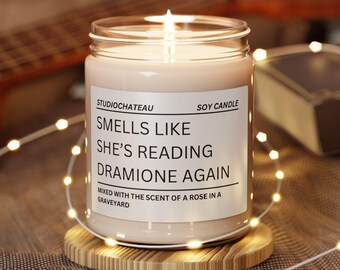 May include: A soy candle in a clear glass jar with a white label that reads "Studio Chateau Soy Candle Smells Like She's Reading Dramione Again Mixed with the scent of a rose in a graveyard".