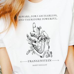 May include: White t-shirt with a black and white illustration of a hand holding a heart, adorned with botanical details. The shirt displays the quote "BEWARE; FOR I AM FEARLESS, AND THEREFORE POWERFUL" and the text "FRANKENSTEIN MARY SHELLEY".