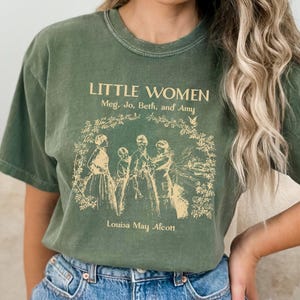 May include: A green t-shirt featuring a vintage-style graphic of the book cover for "Little Women" by Louisa May Alcott. The graphic depicts four women in a garden setting with the text "Little Women, Meg, Jo, Beth, and Amy".
