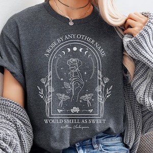 May include: A dark gray t-shirt with a white graphic design featuring a hand holding a rose with a crescent moon and stars in the background. The text "A rose by any other name would smell as sweet" is printed below the graphic. The quote is attributed to William Shakespeare.