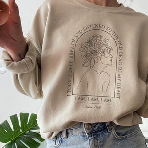 Peut inclure: Un sweat-shirt beige avec un imprimé graphique d'une femme avec des fleurs dans les cheveux. Le texte sur le sweat-shirt est "I took a deep breath and listened to the old brag of my heart. I am, I am, I am. Sylvia Plath."