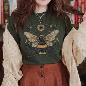 May include: A dark green t-shirt with a stylized illustration of a bee with yellow and black stripes surrounded by flowers. The bee is centered on the shirt and the flowers are arranged around it. The shirt is being worn by a person with long brown hair and a brown skirt.