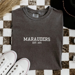 May include: A dark gray t-shirt with the word "MARAUDERS" and "EST. 1971" printed in white. The shirt is laid flat with a pair of white sneakers, a black hat, and decorative disco balls on a checkered surface.