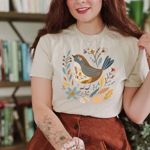 May include: A woman wearing a beige t-shirt with a colorful illustration of a bird and floral design. The shirt has a button closure at the neck. She is wearing a brown skirt and has a book in her hand.
