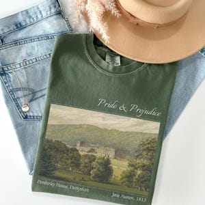 Puede incluir: Una camiseta verde oliva con el texto "Pride & Prejudice" y una imagen de paisaje de Pemberley House, Derbyshire. La camiseta se combina con unos vaqueros de lavado claro, un collar dorado y un sombrero beige con una banda marrón.