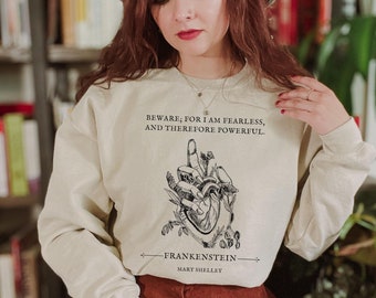 May include: A beige sweatshirt with a black and white illustration of a hand holding a heart. The text "BEWARE, FOR I AM FEARLESS, AND THEREFORE POWERFUL." is printed on the sweatshirt. The text "FRANKENSTEIN" and "MARY SHELLEY" are printed below the illustration.