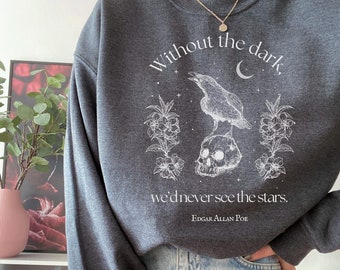 Può includere: Una felpa grigia scura con una stampa grafica bianca di un corvo appollaiato su uno scheletro con fiori e una luna crescente. Il testo recita "Without the dark, we'd never see the stars. Edgar Allan Poe".