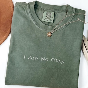 Może przedstawiać: Oliwkowo-zielona koszulka z haftowanym napisem "I am No man". Złoty naszyjnik z wisiorkiem spoczywa na koszulce. Koszulka jest złożona, a na metce widnieje napis "Comfort Colors".