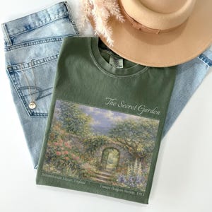 May include: An olive green t-shirt with a landscape print of a garden scene, the text "The Secret Garden", and light blue jeans. A tan hat and a gold necklace are also in the image.