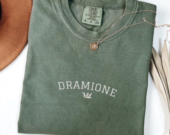 Puede incluir: Una camiseta verde con el texto "DRAMIONE" y un gráfico de corona impreso en la parte delantera. La camiseta está hecha de un material suave y cómodo.