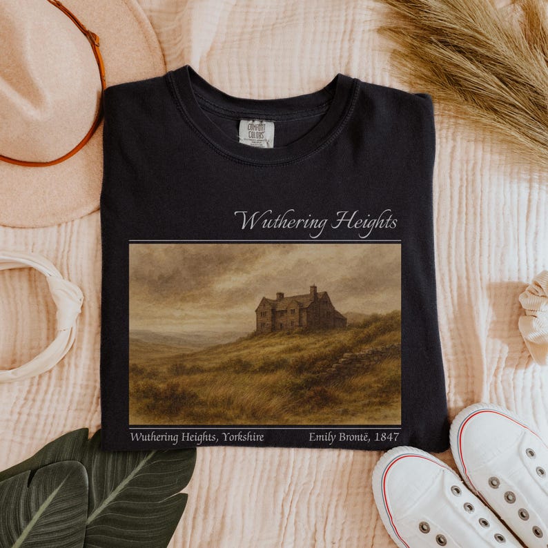 Op de afbeelding: Zwart T-shirt met een landschapsafbeelding van Wuthering Heights. De afbeelding toont een huis op een heuvel, met de tekst "Wuthering Heights" erboven en "Wuthering Heights, Yorkshire Emily Bront&euml;, 1847" eronder.