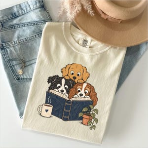 Op de afbeelding: Een crèmekleurig t-shirt met een afbeelding van drie honden die in een donkerblauw boek lezen. Eén hond is goudkleurig, één is zwart-wit en één is bruin-wit. Een klein kopje met een hartje en een plant in een pot staan naast het boek.